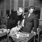 Biskop John Cullberg är på besök i den nyöppnade Gäddgårdsskolan. Lärarinnan Karin Eriksson och hennes klass visar sin verksamhet. Just nu är det teckningslektion. En pojke målar med vattenfärger.