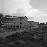 Gäddgårdsskolan börjar bli färdig att tas i bruk. Fönstren är öppna för vädring. Det ligger virke på gården, en cementblandare står kvar. Män cyklar. En bil står parkerad utanför huset.