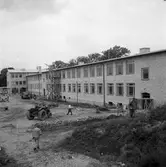 Gäddgårdsskolan byggs. Marken, framför byggnaden, är inte färdigställd. Flera män är i arbete. En byggnadsställning står rest mot husväggen. En lastbil och en traktor används i arbetet. Långt bort, till vänster, står en cementblandare. Till höger går en man med en lie.