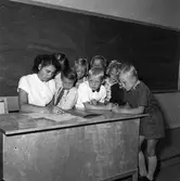 Lärarinnan Gun Martinsson sitter vid katedern omgiven av sina elever. Pojken i vit skjorta är Roland Andersson. Längst till höger står Sivert Karlsson. 
Det är första terminen i den nybyggda Gäddgårdsskolan.