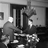 Hantverksföreningens ordförande, Gustaf Johansson (Murar-Gustaf), delar ut mästarbrev i teoretisk och praktisk optik till Sune Gerhardsson.
Platsen är kommunfullmäktigesalen på Rådhuset.