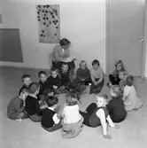 HSB´s barnträdgård. Fröken sitter på en stol och läser i en bok. Barnen sitter i ring på golvet. Mitt i ringen står en liten julgran.
(HSB betyder Hyresgästernas sparkasse- och byggnadsförening)