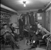 Rökpaus i HSB´s verkstadslokaler. Mannen längst till höger heter Erik Pehrsson/Persson.
Sex män har rast i källarlokalen. Fyra röker pipa, en cigarett och är passiv rökare. De har arbetskläder på sig, skyddsrockar och blåkläder. Efter ena väggen har de glasburkar med skruvar och andra småprylar i. En hatthylla är full av jackor och mössor. I hörnet finns ett handfat. En naken glödlampa lyser i taket.
(Hyresgästernas sparkasse- och byggnadsförening)