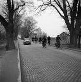 Jädersvägen. Män och kvinnor cyklar till arbetet på Arboga Mekaniska Verkstad. Huset, till vänster, är Gärstadal, bryggeribostäder. Bryggeriet ligger till höger, utanför bilden.
En bil och en moped. Gatsten.
AMV
