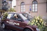Arbogakarnevalen. Paraden har nått Hökenbergsgränd. Här kommer en bil dekorerad med blommor från Lundins blommor. På passagerarsätet sitter Ulla Lundin.
Byggnaden i bakgrunden är Nikolai kulturhus.