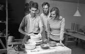 Keramiker i arbete. Ulf Johansson drejar medan Gösta Gräs och Kerstin Hörnlund ser på. I lokalen ses arbetsbord och hyllor med keramik.
Läs Kerstins text 