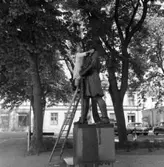 Kyrkvaktmästare Paul Eklund tvättar av Engelbrektsstatyn
Man på stege med vattenhink och borste. Bilar parkerade på Kyrkogatan.