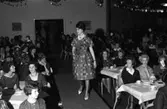Öhrman och Melander har modevisning på Folkan/Medborgarhuset. Personal och medlemmar i Arboga Lottakår ställer upp som mannekänger. Här visar Gunilla Strindler en klänning med skärp i midjan och vidd i kjolen. Publiken sitter vid bord och dricker kaffe.
