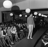 Ungdomsträff med modevisning. Ung kvinna visar polojumper och rutiga långbyxor. Publiken sitter i rader.