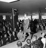 Det är modevisning på Arboga Bil AB. Lokalen är pyntad med björkris och ballonger. Det är möjligen 1-årsjubileum. Bilhallen är full av människor. De flesta har ytterkläder på sig. En ung kvinna, Jakobsson, visar upp en lätt jacka med skärp i midjan. Alldeles nedanför henne sitter Greta Helander med dottern Katarina i knät (flickan har tofsmössa). Till vänster om den närmaste pelaren sitter en kvinna i hatt och glasögon. Det är fröken Eklund som jobbade på Skandinaviska Enskilda Banken.