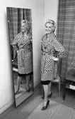 Kvinna i hundtandsmönstrad kappa. Hon står i ett omklädningsrum med spegel. Kvinnan är en av mannekängerna när Öhrman och Melander har sin modevisning på Folkan/Medborgarhuset.