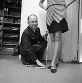 Sture Melander på Öhrman & Melander på Nygatan.
Han sitter på huk, har ett måttband i händerna och mäter klänningslängden åt en kvinna.