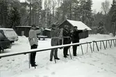 Polisen tränar pistolskytte. Fyra män uppställda. Det snöar.
Ett litet hus och en bil i bakgrunden.