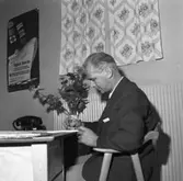 Resebyrån Reso i nya lokaler. En man sitter vid skrivbordet. Han har en penna i handen. En telefon och en vas med blommor står på skrivbordet. På väggen hänger en affisch som lockar med resor till Amerika.