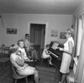 Musiklektion för funktionshindrade elever. Lärarinnan dirigerar gruppen med rytminstrument. En liten flicka spelar piano. Mannen närmast dörren heter Börje Roslund.
Plantsen är möjligen Meijerska gården.