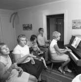 Musiklektion för funktionshindrade. En kvinna spelar piano och eleverna kompar med rytminstrument. Vid dörröppningen sitter Börje Roslund.
Platsen är troligen Mejerska gården.