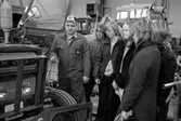 En skolklass är på studiebesök på Nerikes/Närkes Maskiner. Sven Karlsson, i keps och overall, berättar om traktorn. Intill honom står Roger Hedenström, Björn Asp, Lena Oscarsson och Viola Karlsson.