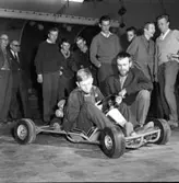 Speed-Car. Män, i olika åldrar, samlade runt en pojke som sitter i en gokart.