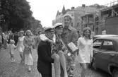 Studentdag! Kicki Holm, iklädd studentmössa och blombuketter, bärs av Anders Holm (i mössa) och Bo Pilerud.
Det är mycket folk i rörelse på Järntorget.