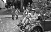 Studentdag! Anders Hellström körs runt i framluckan på en lövad Folkvagn. Anders har studentmössan på sig och blommor i knät.
I bakgrunden bär tre pojkar på sina blåsinstrument. De tillhör ungdomsmusikkåren och har avslutat sitt uppdrag.