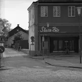 Biografen Sture-Bio, i Lundbergska fastigheten, i hörnet Stora torget - Västerlånggatan. På biografens trappa sitter en ung man. En person cyklar på Västerlånggatan. Trafikskyltar.
