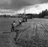 Sturevallen. Här förbereds för en ny ishockeybana. Två män och en traktor i arbete. Mannen i förgrunden är Olle Kropp.
Bakom läktaren anas Arboga Margarinfabrik och tornet på Sankt Nikolai kyrka.