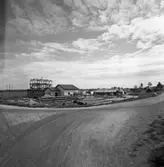 Sågen, sågverket, exteriör.
Timmer ligger utanför. En grusväg i förgrunden. Husen, på kullen, ligger efter Prästgatan.
Till vänster en transformatorstation.