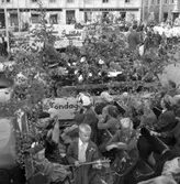 Söndagsskolans Dag firas. Flera lastbilar, med lövade flak, står uppställda på Stora torget. På flaken sitter barn men två lastbilar transporterar var sin blåsorkester. I bakgrunden ses butikerna Haglund och Eklöws Bosättning.