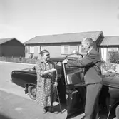 En kvinna ska åka taxi. Hon har en käpp i handen. Den uniformerade chauffören håller upp bildörren.