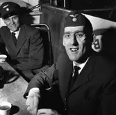 Arne Fallkvist (närmast kameran) och Karl-Erik Larsson har kafferast på taxistationen. Arne, som är en duktig ishockeyspelare, har ett plåster under ögat. De har var sin kopp kaffe framför sig på bordet.
Arne har båtmössa och Karl-Erik har skärmmössa.