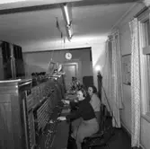 Telefonister på Televerket i Sohlénska huset på Nygatan. Kvinnorna arbetar vid växelbordet. Var och en har en telefonlur intill örat.
En klocka hänger på väggen ovanför dörren.
Läs om Telefonen i Arboga och branden på Televerket 1956 i Arboga Minnes årsbok 1993.
Läs om Televerket och branden 1956 i Arboga Minnes årsbok 1993.