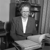 Telekommissarie Eva Geete på Televerket, Sohlénska huset, på Nygatan.
Eva bär glasögon och är klädd i en dräktjacka med en brosch på slaget.
Läs om Televerket och branden 1956 i Arboga Minnes årsbok 1993.
Kvinna som sitter vid skrivbordet och bläddrar i en pärm. Bakom henne ses en skrivmaskin på ett lågt bord, en bokhylla med pärmar samt en gardin.