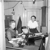 Televerket. Arbetet fortsätter trots skadorna efter branden i fastigheten på Nygatan.
En kvinna sitter vid ett skrivbord. Hon har en pärm uppslagen. En telefon står på bordet. En annan kvinna står bredvid skrivbordet. En lampsladd hänger i taket.
Läs om Telefonen i Arboga och branden på Televerket 1956 i Arboga Minnes årsbok 1993.