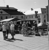 Torghandel. Nanny och Göte Lundin säljer blommor på Stora torget. De har sin skåpbil med sig. Flera kvinnor har samlats vid deras torgstånd. Kvinnan som håller i krukväxten kallas för 