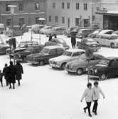 Bilar parkerade på Stora torget. Det är vinter och snö. Människor promenerar. Stadsgården och Sture-bio ses i bakgrunden.
Bland bilarna ses SAAB, Volvo PV och Volvo Amazon.