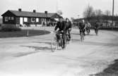 Män som cyklar hem efter arbetsdagens slut, på CVA, Centrala Verkstaden Arboga. I bakgrunden ses människor på väg till bilparkeringen eller busshållplatsen. I huset sitter 