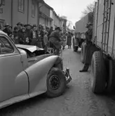 Trafikolycka. En personbil har krockat med ett lastbilssläp. Motorhuven och stötfångaren är skadade. Många människor har samlats för att se på. Till höger kommer en man med kamera.