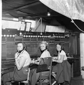 Trots skadorna efter branden, fortsätter arbetet på Televerket på Nygatan. Tre kvinnor som arbetar vid växelbordet. De har varsin telefonlur vid örat.
Från vänster: Kerstin Örnefur, Jansson (gift med frisör Folke Jansson med salong på Paradisgränd), okänd
Det hänger en presenning över arbetsplatsen. En naken glödlampa hänger i taket.
Läs om Telefonen i Arboga och branden på Televerket 1956 i Arboga Minnes årsbok 1993.