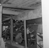 Trots branden i Televerkets hus på Nygatan, fortsätter kvinnorna i växeln sitt arbete. Växeltelefonisterna sitter vid växelbord. De har en telefonlur vid örat. Män, i arbetskläder, inspekterar brandskadorna. En presenning är spänd över arbetsplatsen.
Läs om Telefonen i Arboga och branden på Televerket 1956 i Arboga Minnes årsbok 1993.