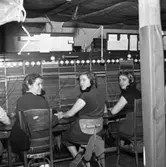 Trots brandskadorna i Televerket på Nygatan, så fortsätter telefonisterna arbetet vid växelborden. I mitten sitter Mary Pettersson. Kvinnorna har en telefonlur fastsatt vid vänster öra. En presenning är spänd över arbetsplatsen för att inte vattnet, från släckningsarbetet, ska skada växeln. En ensam glödlampa hänger i taket.
Läs om Telefonen i Arboga och branden på Televerket 1956 i Arboga Minnes årsbok 1993.