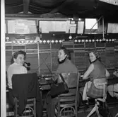 Telefonisterna fortsätter sitt arbete vid växelborden trots skadorna efter branden i fastigheten. En presenning är spänd över arbetsplatsen för att inte vatten, från släckningsarbetet, ska skada växeln.
Klockorna, som hänger på rad, visar hur många perioder samtalen varat. Kvinnorna har en telefonlur hängande intill örat.
Från vänster: okänd, Alice Ståhl och Britt Johansson.
Televerket låg på Nygatan.
Läs om Telefonen i Arboga och branden på Televerket 1956 i Arboga Minnes årsbok 1993.