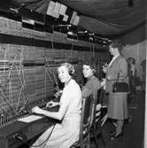 Televerket på Nygatan. Trots skadorna efter branden fortsätter arbetet vid växelborden.
Växeltelefonisterna sitter på rad. Var och en har en telefonlur vid sitt vänstra öra. Bakom kvinnorna står telekommissarie Eva Geete/Gethe.
I taket hänger en presenning som skydd mot det vatten som användes för att bekämta branden.
Läs om Telefonen i Arboga och branden på Televerket 1956 i Arboga Minnes årsbok 1993.