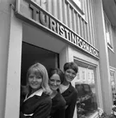 Turistbyrån stänger för säsongen. Tre turistvärdinnor i sina uniformer. Souvernirer visas i skyltfönstret. En balkonglåda, med blommor, sitter på husväggen.
Troligen Västerlånggatan.