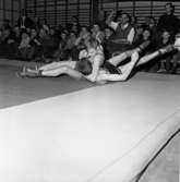 Ungdomsbrottning. Två brottare på mattan. Matchen äger rum i en gymnastiksal. Publiken sitter nedanför ribbstolarna.