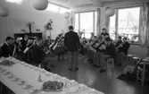 Ungdomsorkestern/blåsorkestern uppträder. Dirigent och musiker är klädda i uniform. Kaffebordet står dukat med bullar på faten.