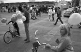 Valmöte på Nytorget. Grillen och Pressbyrån ses i bakgrunden. Folkvagnen svänger västerut, på Ahllöfsgatan.
Mannen med ballongerna är Per-Olov Nilsson. Det är mycket folk i rörelse på torget. En del har paraply. I förgrunden ses några barn med cyklar.