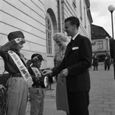 På väg till valurnan stannar det nyförlovade paret, Sigbritt och Yngvar Gunnarsson, och skänker en slant till insamlingen som scouterna gör. Den lille pojken med bössan är Rolf Mattsson.
I bakgrunden ses skylten till polisstationen och Normans. Platsen är Rådhusgatan.