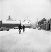 Vintermotiv. Två personer cyklar, i snön, på Östra Hamngatan. En bil kommer åkande. Till vänster ligger Hamntorget, där står ett par bilar parkerade. Till höger skymtar Texacomacken. I bakgrunden ses skorstenen på Arboga Kvarn och Maltfabrik. Tornet på Heliga Trefaldighetskyrkan anas till höger.