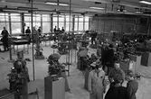 Yrkesskolan, maskinhallen. Elever vid maskiner. Två lärare, iklädda skyddsrockar, ses i bildens nedre högra hörn. Den vänstra, av dem, är yrkeslärare Otto Daunfelt. En man i hatt och ytterrock är på besök, mitt i bilden.
Bilden ser ut att vara tagen i verkstaden på Vasagymnasiet.