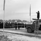Örebrovägen ska öppnas efter ombyggnad. En stor publik vinterklädda människor har samlats vid bandet som snart ska klippas. På taket till en lastbil står en man och håller tal i en megafon. En stege är lutad mot lastbilshytten. På dörren står det 